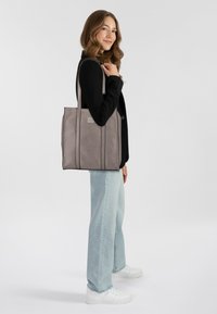 Sac tote en matériau texturé gris avec des accents noirs, forme rectangulaire, deux poignées, fond plat et une petite étiquette à l'avant.