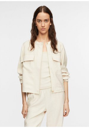 Frau mit langen welligen braunen Haaren trägt eine cremefarbene Jacke, passendes Oberteil und lockere Hose, stehend vor einem schlichten weißen Hintergrund.