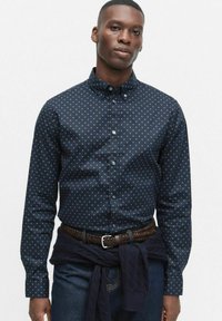 Marinblå skjorta med knappar och ett litet guldstjärnmönster, skräddarsydd passform, långa ärmar och knäppta manschetter, bärs med ett flätat brunt bälte.