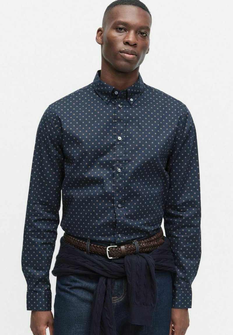 Marinblå skjorta med knappar och ett litet guldstjärnmönster, skräddarsydd passform, långa ärmar och knäppta manschetter, bärs med ett flätat brunt bälte.