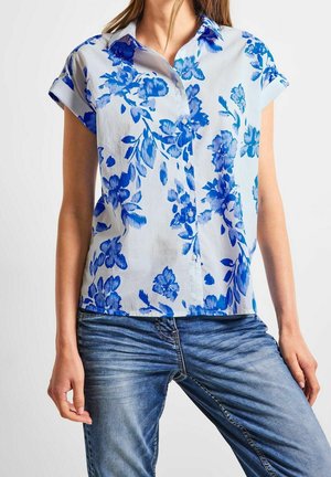 Femme portant un chemisier blanc à manches courtes avec un imprimé floral bleu et un jean en denim bleu, debout devant un fond clair uni.