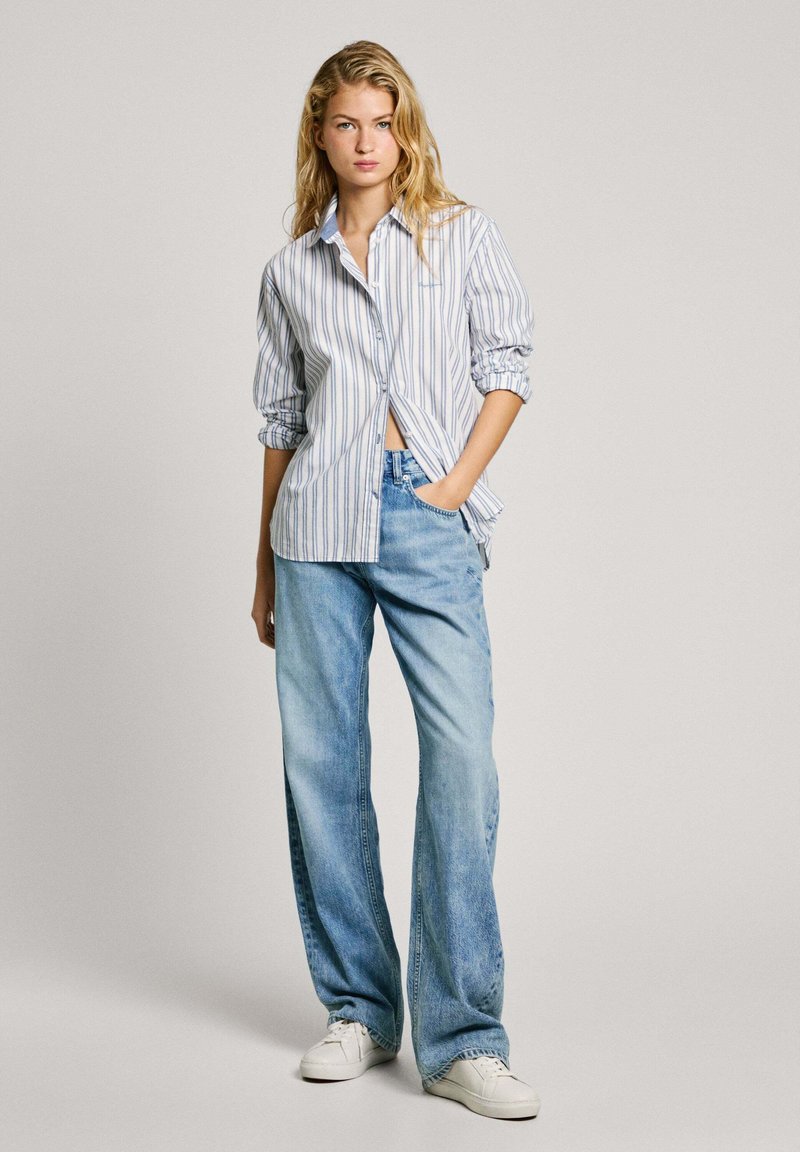 Camicia a righe azzurro e bianco, abbinata a jeans in denim blu a gamba larga e sneakers bianche. Look casual e rilassato, con maniche arrotolate.