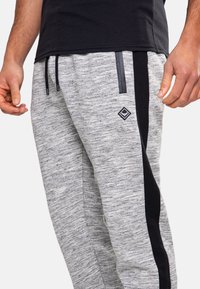 Graue Jogginghosen mit strukturierter Webart, ausgestattet mit einem schwarzen Seitenstreifen, elastischem Bund und schwarzen Kordeln. Kleines Logo-Patch am Oberschenkel.