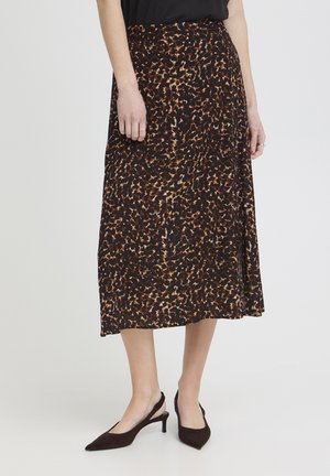 Ženska v midi krilu z rjavim, črnim in bež živalskim potiskom, obute v črne čevlje z odprtim petnim delom in koničastim prstom, stoji ob belem ozadju.