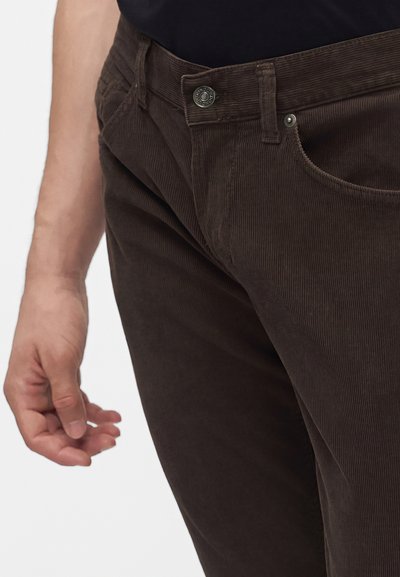 Gros plan d'une personne portant un pantalon en velours côtelé brun foncé et une chemise noire, montrant la poche avant et la zone des boutons.