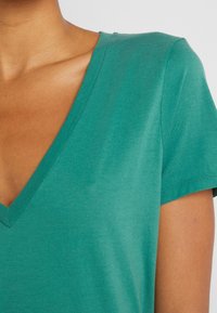 Primo piano di una persona che indossa una maglietta a maniche corte e scollo a V color teal, che mostra la pelle del collo, della spalla e della parte superiore del petto.