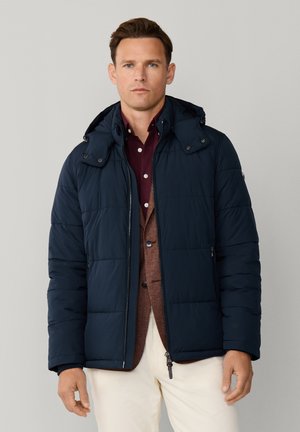 Muž v tmavomodrej prešívanej bunde cez hnedé sako a bordovú košeľu, stojaci pred jednoduchým svetlošedým pozadím.