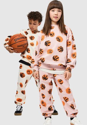 Dos niños llevan conjuntos de ropa cómoda a juego con estampados de balones de baloncesto naranjas y caras sonrientes. La tela es suave y texturizada, en tonos pastel.
