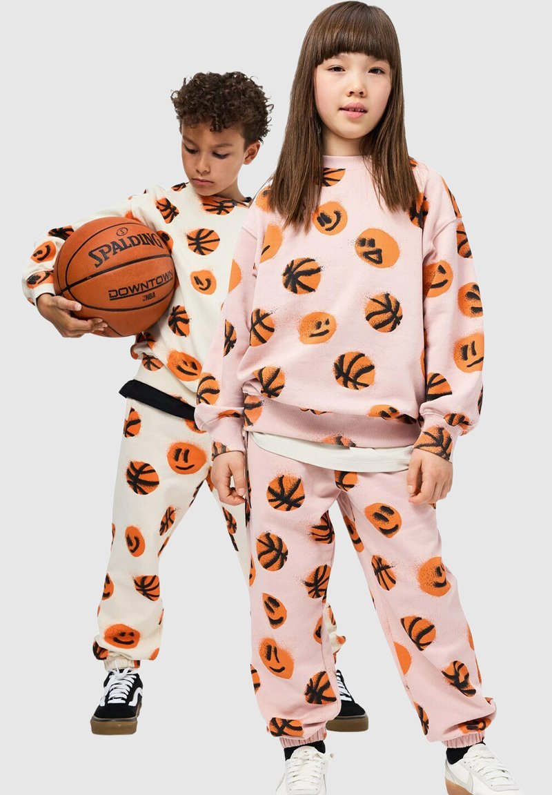 Deux enfants portent des ensembles de vêtements d'intérieur assortis avec des motifs de ballons de basket orange et de visages souriants. Le tissu est doux et texturé, dans des tons pastel.