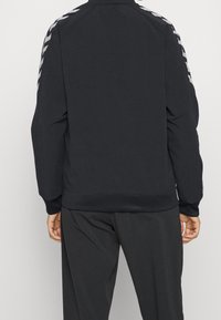Chaqueta deportiva negra con cuello alto, puños acanalados y un acabado texturizado. Presenta rayas en forma de chevron blancas contrastantes en las mangas.