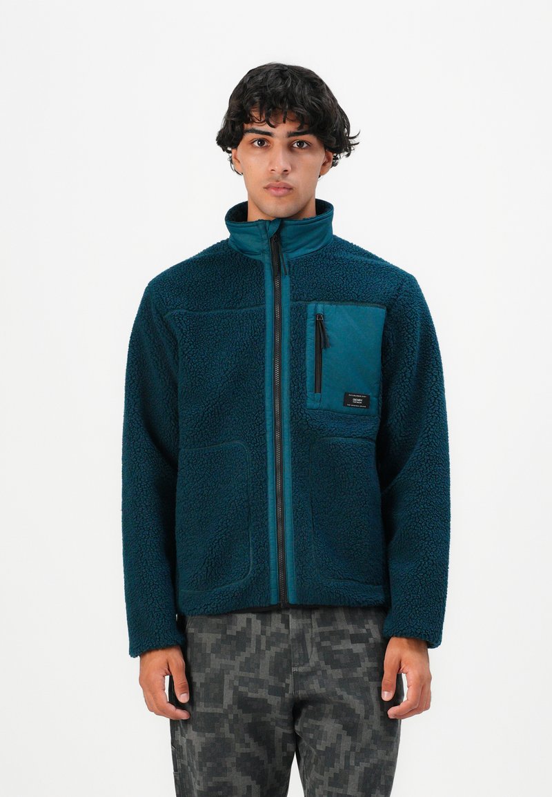 Uomo con capelli ricci scuri che indossa una giacca in fleece teal con tasca frontale zip e pantaloni in camouflage digitale grigi, in piedi davanti a uno sfondo bianco.