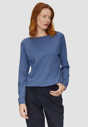 Blue long-sleeve top with a wide neck and ribbed cuffs, made of textured fabric. Worn with dark cargo pants.