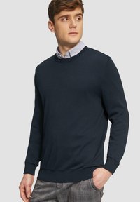 Marine blauwe crewneck trui van zacht materiaal, met lange mouwen en ribgebreide manchetten. Draag over een licht overhemd met kraag, gecombineerd met een geruite broek.
