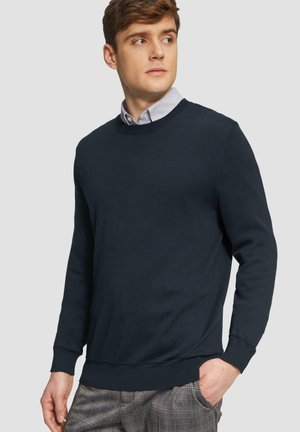 Marinblauer Rundhalsausschnitt-Pullover aus weichem Material, mit langen Ärmeln und gerippten Bündchen. Über einem hellen Hemd mit Kragen getragen, kombiniert mit karierter Hose.