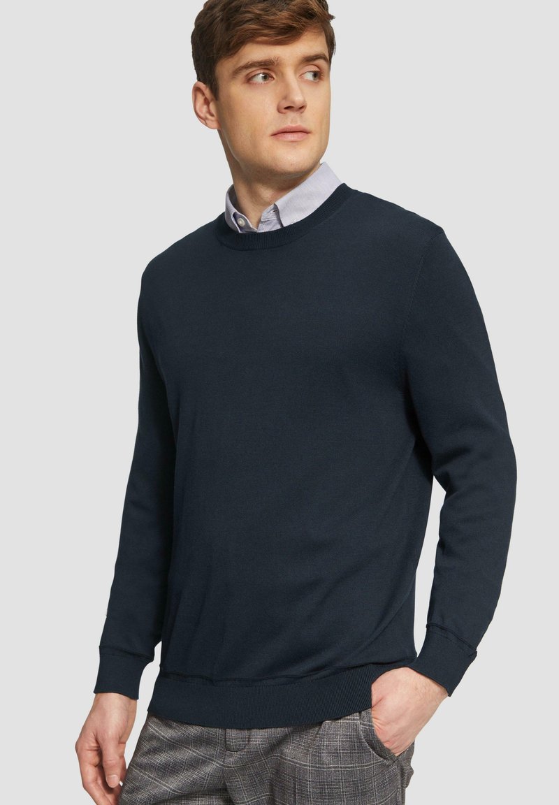Marine blauwe crewneck trui van zacht materiaal, met lange mouwen en ribgebreide manchetten. Draag over een licht overhemd met kraag, gecombineerd met een geruite broek.