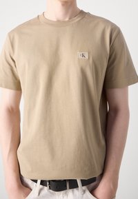 Tan bomulls-t-shirt med ikonisk logotyp på bröstet, kortärmad och med rundad halsringning, med en mjuk textur.