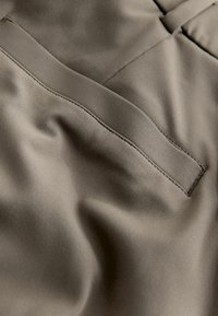 Sileä kangas hillityssä oliivinvihreässä, jossa on hienovarainen tekstuuri ja ommeltu yksityiskohta. Kangas lähikuvassa paljastaa pehmeät laskokset ja taskun.