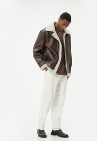 Giacca di pelle marrone con colletto in shearling, t-shirt marrone, pantaloni cargo bianchi e stivali scuri. L'outfit presenta linee pulite e trame a strati.
