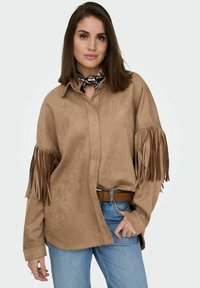 Tanfarvet ruskinds skjorte med lange frynser på ærmerne. Båret over lyseblå jeans og kombineret med et brunt bælte. Tørklæde omkring halsen tilføjer mønster.