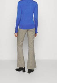 Blauer Strickpullover mit taillierter Passform und langen Ärmeln, die mit Knopfakzenten versehen sind. Kombiniert mit ausgestellten Hosen im Hahnentrittmuster und schwarzen High Heels.