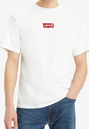Homme portant un t-shirt Levi's blanc avec un logo rouge sur la poitrine et un jean bleu, main dans la poche, fond uni.