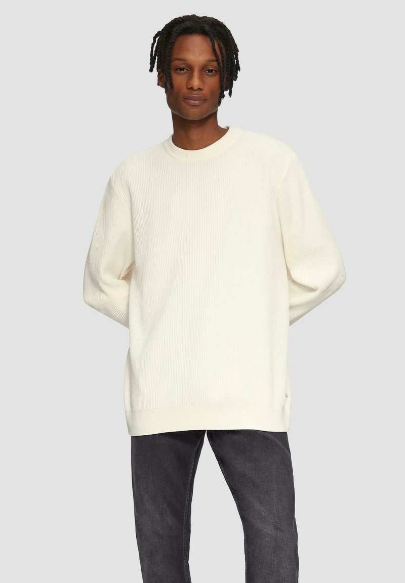 Off-white, geripptes Sweater mit rundem Ausschnitt, überschnittenen Schultern und entspannter Passform, kombiniert mit dunkelgrauen Jeans.