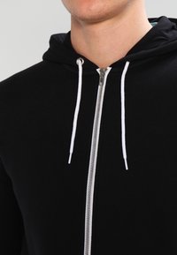 Sudadera con cremallera negra y cremallera blanca, con cordones, que presenta una textura suave y capucha, diseñada para uso casual.