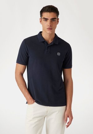 Camisa polo de color navy hecha de algodón, con una textura suave, mangas cortas y un parche de la marca en el lado izquierdo del pecho. Combinada con jeans claros.