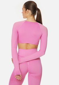 Prilega majica s dolgimi rokavi v svetlo rožnati barvi, z gladko teksturo in raglan rokavi, dopolnjena z ujemajočimi se visoko pasnimi pajkicami.