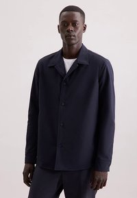 Granatowa kurtka na guziki z gładkiej tkaniny z szpiczastym kołnierzem, długimi rękawami i subtelnymi detalami szwów. Prosty design, minimalistyczne akcenty.