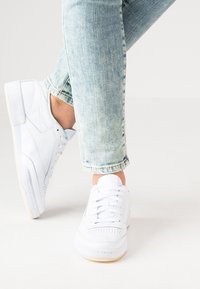 Vita lädersneakers med perforerade detaljer, runda snörningar och en texturerad gummisula. Bärs med ljusblå, något slitna jeans.