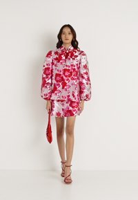 Ρουχισμός με λουλουδάτο μοτίβο σε λαμπερό ροζ και κόκκινο ύφασμα; περιλαμβάνει ψηλό γιακά, φουσκωτούς ώμους και κοντή φούστα, συνδυασμένο με μια κόκκινη τσάντα.