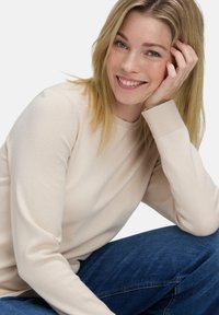 Mosolygó szőke nő a kezére támaszkodva, bézs pulóvert és kék farmernadrágot viselve, egy sima fehér háttér előtt.