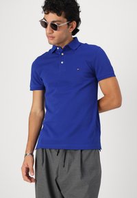 Polo de algodón azul con cuello, dos botones y detalle de logo. Combinado con pantalones grises con cordón. El modelo lleva gafas de sol y tiene el cabello rizado.