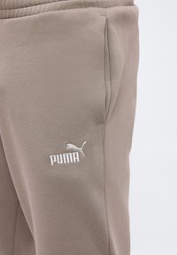 Κοντινή λήψη μπεζ παντελονιών Puma που δείχνει πλαϊνή τσέπη και λευκό κεντημένο λογότυπο Puma στο μηρό.