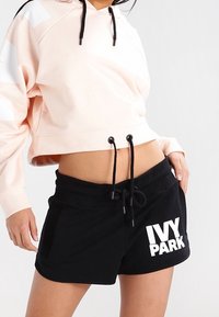 Cropped hoodie rosa chiaro con coulisse nere e dettagli bianchi, abbinato a pantaloni corti neri con la scritta "IVY PARK" in grassetto bianco.