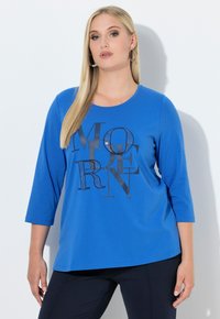 Niebieska koszulka z długim rękawem i okrągłym dekoltem, ozdobiona czarnym, błyszczącym napisem. Miękka tkanina, luźny krój, zestawiona z ciemnymi spodniami.