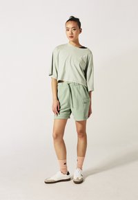 T-shirt di cotone morbido, verde chiaro e corto, abbinato a pantaloni corti coordinati. L'outfit presenta un cordoncino e una tasca laterale. Sneakers bianche con calzini rosa.