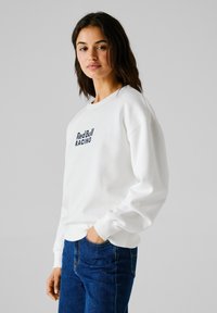 Sudadera blanca con cuello redondo y puños de canalé, con "Red Bull RACING" impreso en letras marinas en el frente. Combinada con jeans azules.