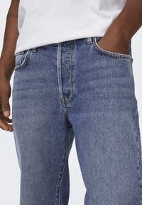 Jean en denim bleu avec un design à jambe droite, doté d'une taille mi-haute, de cinq poches et de coutures jaunes contrastées. Texture lisse.
