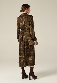 Abito lungo marrone con motivo circolare, vita stretta, maniche lunghe e gonna fluida. Abbinato a tacchi a piattaforma e una pochette marrone.