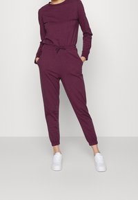 Completo jogger color marsala realizzato in tessuto morbido; maglia a maniche lunghe con scollatura tonda, vita elasticizzata e gambe affusolate; abbinato a sneakers bianche.
