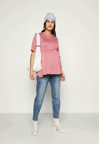 Blusa de maternidade rosa com mangas curtas, detalhe de cintura franzida, combinada com jeans azul claro e botins brancos. Acessórios incluem um gorro azul claro e uma mala tote branca.