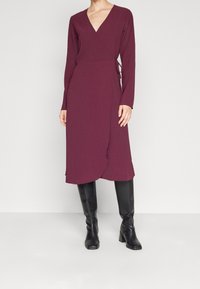 Kvinna klädd i en långärmad, vinröd omlottkjol i kombination med höga svarta läderstövlar, stående mot en enkel ljus bakgrund.