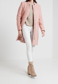 Abrigo texturizado rosa con corte recto, cierres a presión, llevado sobre un suéter beige y jeans ajustados blancos, combinado con botas de tacón beige.