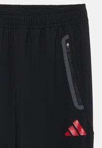 Čierne športové nohavice s elastickým pásom, bočnou zipsovou kapsou, sivými akcentmi a červeným logom Adidas na dolnej ľavej strane.