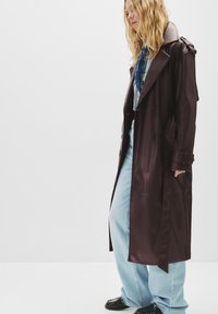 Cappotto trench in pelle marrone con ampio colletto, design doppiopetto e maniche con cintura, abbinato a jeans larghi di colore blu chiaro e scarpe nere.