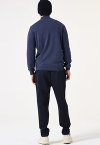 Marineblauwe trui met een gestructureerde afwerking, geribbelde manchetten, gedragen met donkere broek en een gebreide muts, gezien van achteren.