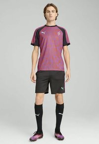 Fiatal férfi lila és narancssárga mintás sportmezben, fekete rövidnadrágban, fekete térdig érő zokniban és rózsaszín focicipőben áll.