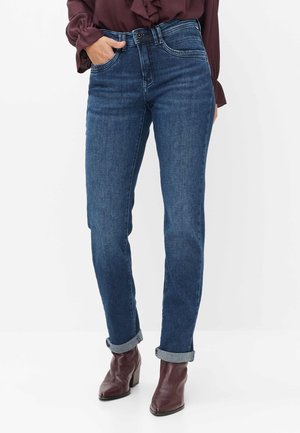 Frau trägt dunkelblaue hochgekrempelte Jeans, braune Stiefeletten und eine burgunderfarbene Langarmbluse mit Rüschen vorne.